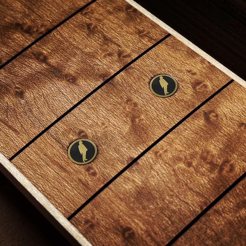 May include: A close-up shot of a wooden surface with a warm brown hue. The wood grain is visible, with dark lines dividing the panels. Two circular, gold-edged black emblems, each featuring a person's silhouette, are present.