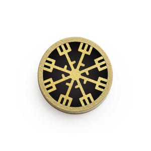 May include: A round, gold-colored pendant with a black center. The pendant features a gold-colored, stylized snowflake design. The pendant is set against a white background.