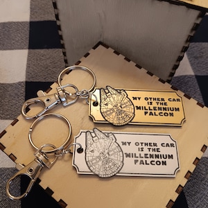 Puede incluir: Dos llaveros plateados con una etiqueta de madera unida a cada uno. Las etiquetas son rectangulares y tienen una imagen en blanco y negro de la nave espacial Millennium Falcon. El texto en las etiquetas dice "Mi otro coche es el Millennium Falcon".