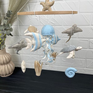 May include: A baby mobile with a wooden frame and felt sea creatures, including a blue jellyfish, a grey turtle, a grey shark, a blue and white fish, a grey stingray, a starfish, a seashell, and two seagrass plants.