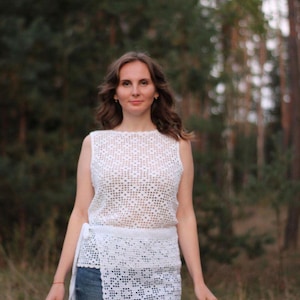 May include: A white, sleeveless crochet top and matching wrap skirt. The top and skirt are made of a delicate, open-weave pattern. The skirt is wrapped around the waist and tied with a long sash. The person is wearing blue jeans.