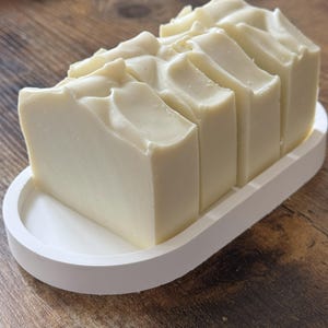 Puede incluir: Un bloque rectangular de jabón de color claro, cortado en barras individuales, descansa sobre una jabonera blanca de forma ovalada. El jabón tiene una superficie texturizada, y la jabonera está sobre una superficie de madera.