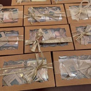 May include: Several brown cardboard boxes with clear window tops are arranged in a group. Each box is tied with a gold ribbon. The boxes contain various items, including soaps and small cards. The overall theme is gift sets.
