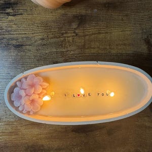 May include: A peach-colored, oval-shaped candle with the words "I love you" spelled out in small letters. Pink flower accents are on the left side. The candle is in a white tray, and the background is a wooden surface.