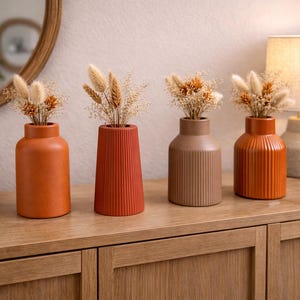 May include: Four ceramic vases in shades of orange and brown, each holding dried floral arrangements. The vases have different shapes and textures, including ribbed and cylindrical designs. The arrangement sits on a wooden cabinet.