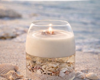 Op de afbeelding: Een brandende, handgemaakte kustkaars in een heldere glazen pot, met een laag zand en schelpen. De kaars staat op een zandstrand met verspreide schelpen. De tekst luidt "Bring the Beach Home".