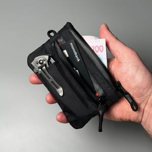 Könnte beinhalten: Schwarzes Portemonnaie mit Reißverschlussfach, das eine Kreditkarte mit der Aufschrift "monobank" und ein Taschenmesser enthält. Das Portemonnaie enthält auch Papiergeld. Das Portemonnaie wird in einer Hand gehalten.