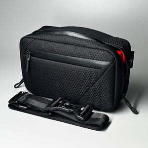 Puede incluir: Un bolso bandolera rectangular negro con un patrón hexagonal texturizado en el exterior, un bolsillo frontal con cremallera horizontal y una correa de nailon negra desmontable con un cierre de hebilla de plástico resistente. Se ve un interior rojo.