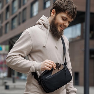 May include: A black crossbody bag with a phone in the front pocket. The bag has a black strap and is worn over the shoulder. The person is wearing a beige hoodie and dark jeans.
