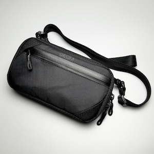 May include: Black crossbody bag with a zippered main compartment and adjustable strap. The bag features the word "GEOM" printed on the front. The bag has a sleek, modern design.
