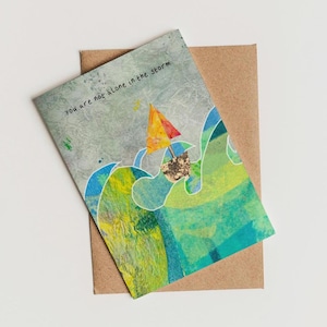 May include: A greeting card featuring a watercolour illustration of a sailboat navigating stormy seas. The card reads "You are not alone in the storm".