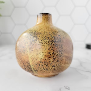 May include: A small, round ceramic vase with a narrow neck. The vase features a speckled, mottled pattern in shades of brown, tan, and black. This decorative vase is likely used for holding flowers or as a standalone accent piece.