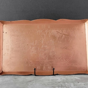 May include: A copper serving tray with an ornate design featuring a scene of people and a landscape. The tray has a scalloped edge and is decorated with Chinese characters.