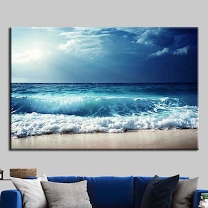 May include: A large canvas print depicting a blue ocean wave crashing on a sandy beach. The sky is a mix of blue and grey with white clouds. The image is framed and hanging on a wall above a blue sofa.