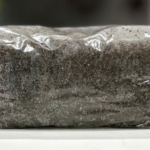 May include: A close-up of a brown, granular substance packaged in a clear plastic bag. The bag is sealed and the substance is visible through the plastic.