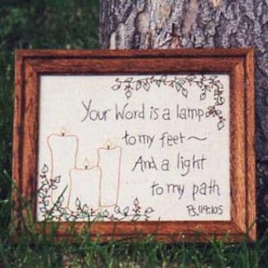 Op de afbeelding: Een ingelijst borduurwerk met een beige achtergrond en een bruine houten lijst. Het borduurwerk toont drie kaarsen met vlammen en de tekst "Your Word is a lamp to my feet~ And a light to my path Ps.119:105".