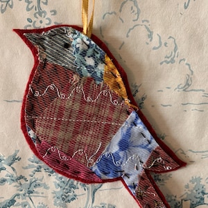 Op de afbeelding: Een rood, blauw en gouden patchwork vogel ornament met een gouden lint eraan bevestigd. De vogel is gemaakt van stofresten en heeft een delicaat, handgestikt ontwerp.