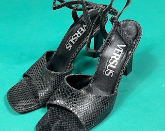 May include: A pair of black snakeskin heeled sandals with ankle straps. The sandals have a square toe and the brand name "VERSUS" is printed on the straps.