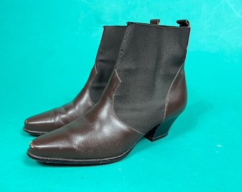 May include: A pair of brown leather ankle boots with a pointed toe and a black elastic panel on the sides. The boots have a small, angled heel. The background is a solid teal colour.
