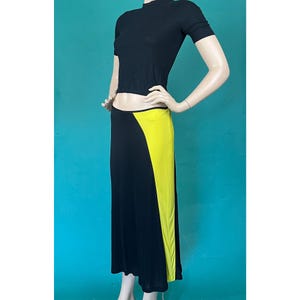 May include: A black, short-sleeved top and a long black skirt with a vibrant yellow panel. The skirt has a high waist and a flowing silhouette. The outfit is displayed on a mannequin against a teal background.