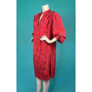 Könnte beinhalten: Ein rotes Kleid mit schwarzem Muster wird auf einer Schaufensterpuppe präsentiert. Das Kleid hat einen Stehkragen, lange Ärmel mit gerafften Schultern und eine Knopfleiste vorne. Der Stoff scheint seidig zu sein.