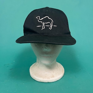 Könnte beinhalten: Eine schwarze Baseballkappe mit einer weißen Stickerei, die den Umriss eines Kamels auf einer horizontalen Linie zeigt. Die Kappe hat eine strukturierte Krone und einen flachen Schirm, mit einem einfachen grafischen Design auf der Vorderseite.