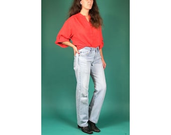 May include: A woman wearing a red button-down shirt and light blue jeans. She is standing in front of a teal background.