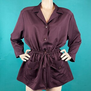 May include: A plum-colored jacket with a collared neckline, button-up front, and drawstring waist. The jacket features two front pockets with button closures and long sleeves. The garment is displayed on a mannequin against a teal background.