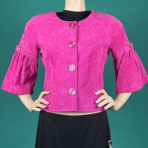 May include: A bright pink suede jacket with three-quarter length sleeves and large buttons down the front. The jacket has a round neckline and flared sleeves. The jacket is displayed on a mannequin.
