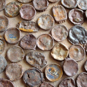 May include: A close-up of a collection of rusty bottle caps, some with faded logos and text, arranged in a pattern on a light brown surface.