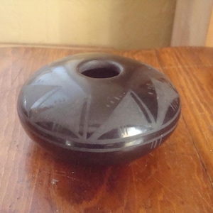 May include: A dark brown, round ceramic vase with a central opening. The vase features etched geometric designs around the top and base. The vase is displayed on a wooden surface.