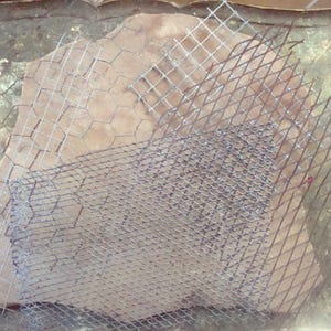 May include: A rough, light brown stone partially covered by two different types of metal mesh. One mesh is a hexagonal chicken wire pattern, and the other is a diamond-shaped expanded metal. The stone has a textured surface.