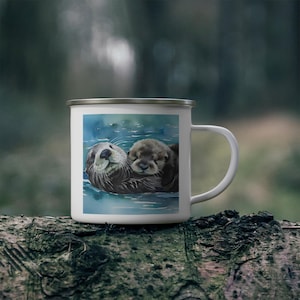 Puede incluir: Taza de esmalte blanco con una ilustración de acuarela de dos nutrias marinas acurrucadas en el agua. Las nutrias son marrones y tienen ojos negros. El agua es azul y verde.