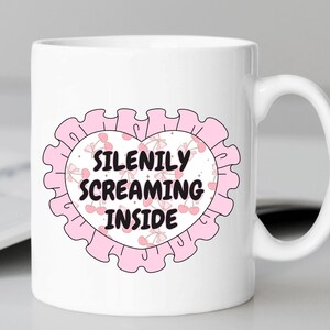 K&ouml;nnte beinhalten: Wei&szlig;e Keramiktasse mit einem rosafarbenen Herzdesign. Das Herz hat einen rosafarbenen R&uuml;schenrand und einen wei&szlig;en Kreis in der Mitte mit dem Text "SILENILY SCREAMING INSIDE" in Schwarz.