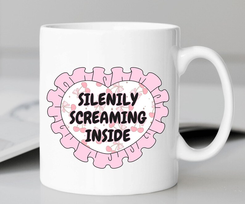 K&ouml;nnte beinhalten: Wei&szlig;e Keramiktasse mit einem rosafarbenen Herzdesign. Das Herz hat einen rosafarbenen R&uuml;schenrand und einen wei&szlig;en Kreis in der Mitte mit dem Text "SILENILY SCREAMING INSIDE" in Schwarz.