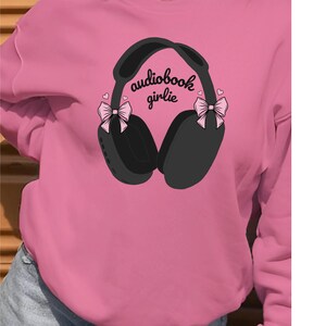 May include: Pink sweatshirt with a graphic of black headphones with pink bows on each side. The text "audiobook girlie" is printed on the headphones.