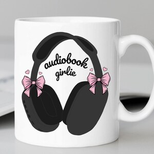 May include: White ceramic mug with a black and white illustration of headphones with pink bows on each side. The text "audiobook girlie" is printed on the mug.