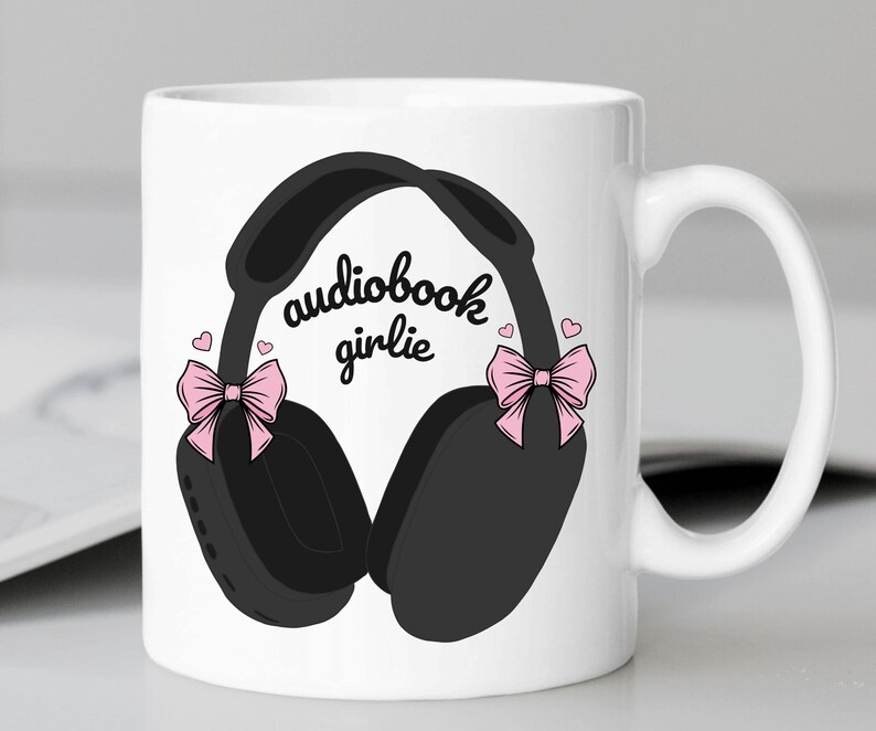 May include: White ceramic mug with a black and white illustration of headphones with pink bows on each side. The text "audiobook girlie" is printed on the mug.