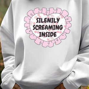 K&ouml;nnte beinhalten: Wei&szlig;er Pullover mit einem rosafarbenen Herzdesign. Das Herz hat einen rosafarbenen gewellten Rand und zeigt den Text "Silently Screaming Inside" mit rosafarbenen Kirschen und Schleifen.