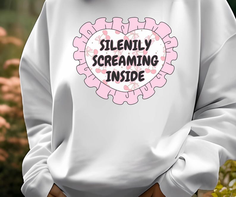 K&ouml;nnte beinhalten: Wei&szlig;er Pullover mit einem rosafarbenen Herzdesign. Das Herz hat einen rosafarbenen gewellten Rand und zeigt den Text "Silently Screaming Inside" mit rosafarbenen Kirschen und Schleifen.