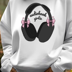 May include: White sweatshirt with a black and gray illustration of headphones with pink bows and the text "audiobook girlie".