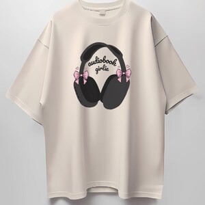May include: A beige t-shirt with a graphic of black headphones with pink bows on each side. The text "audiobook girlie" is printed on the headphones.