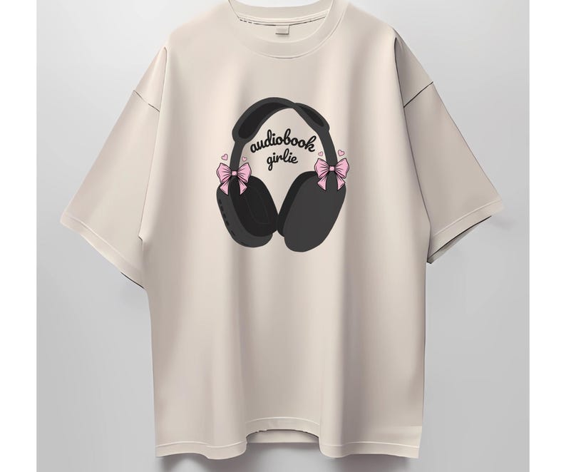 May include: A beige t-shirt with a graphic of black headphones with pink bows on each side. The text "audiobook girlie" is printed on the headphones.
