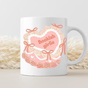 May include: White ceramic mug with a pink and white heart-shaped design. The heart has a white lace trim and the text "Bookish girlie" in orange.
