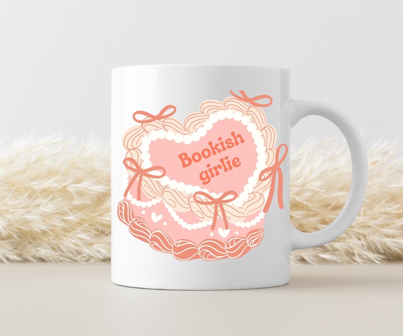 May include: White ceramic mug with a pink and white heart-shaped design. The heart has a white lace trim and the text "Bookish girlie" in orange.