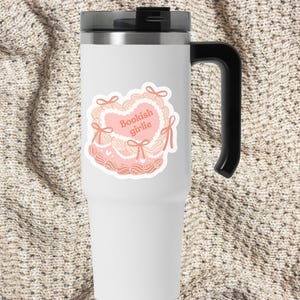May include: White stainless steel travel mug with a black handle. The mug has a pink and white sticker with a heart design and the text "Bookish girlie".