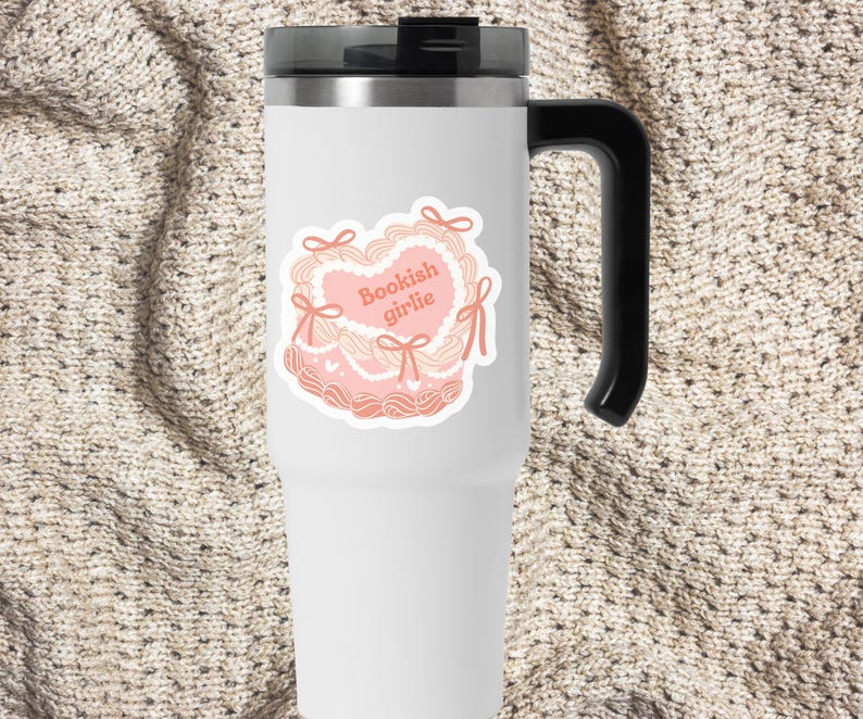 May include: White stainless steel travel mug with a black handle. The mug has a pink and white sticker with a heart design and the text "Bookish girlie".