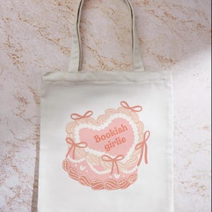 May include: A white canvas tote bag with a pink heart-shaped cake design. The cake has white frosting and pink bows. The text "Bookish girlie" is written in pink on the cake.