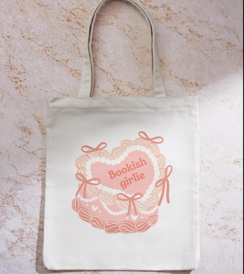 May include: A white canvas tote bag with a pink heart-shaped cake design. The cake has white frosting and pink bows. The text "Bookish girlie" is written in pink on the cake.