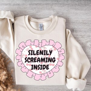 K&ouml;nnte beinhalten: Ein cremefarbener Pullover mit einem rosafarbenen herzf&ouml;rmigen Design. Das Herz hat einen wei&szlig;en Hintergrund mit rosa Kirschen und dem Text "SILENILY SCREAMING INSIDE".